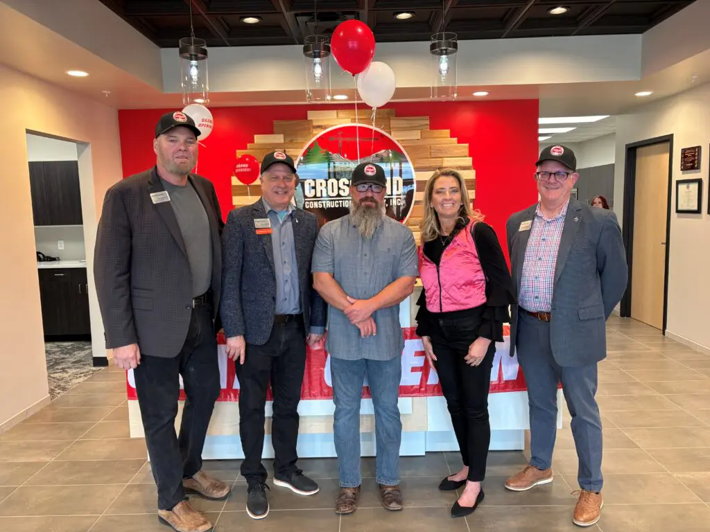 Five adults wearing business attire and baseball caps stand together and smile in a brightly lit office lobby with a red and white wall behind them and balloons above.