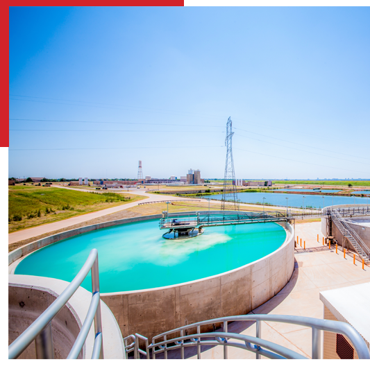 A large circular water treatment tank with turquoise water sits outdoors on a sunny day, surrounded by walkways and railings, with grassy fields and industrial buildings in the background.
