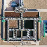 Aerial view of a water treatment facility showing rectangular tanks, walkways, green pipes, and equipment on a concrete and gravel ground.