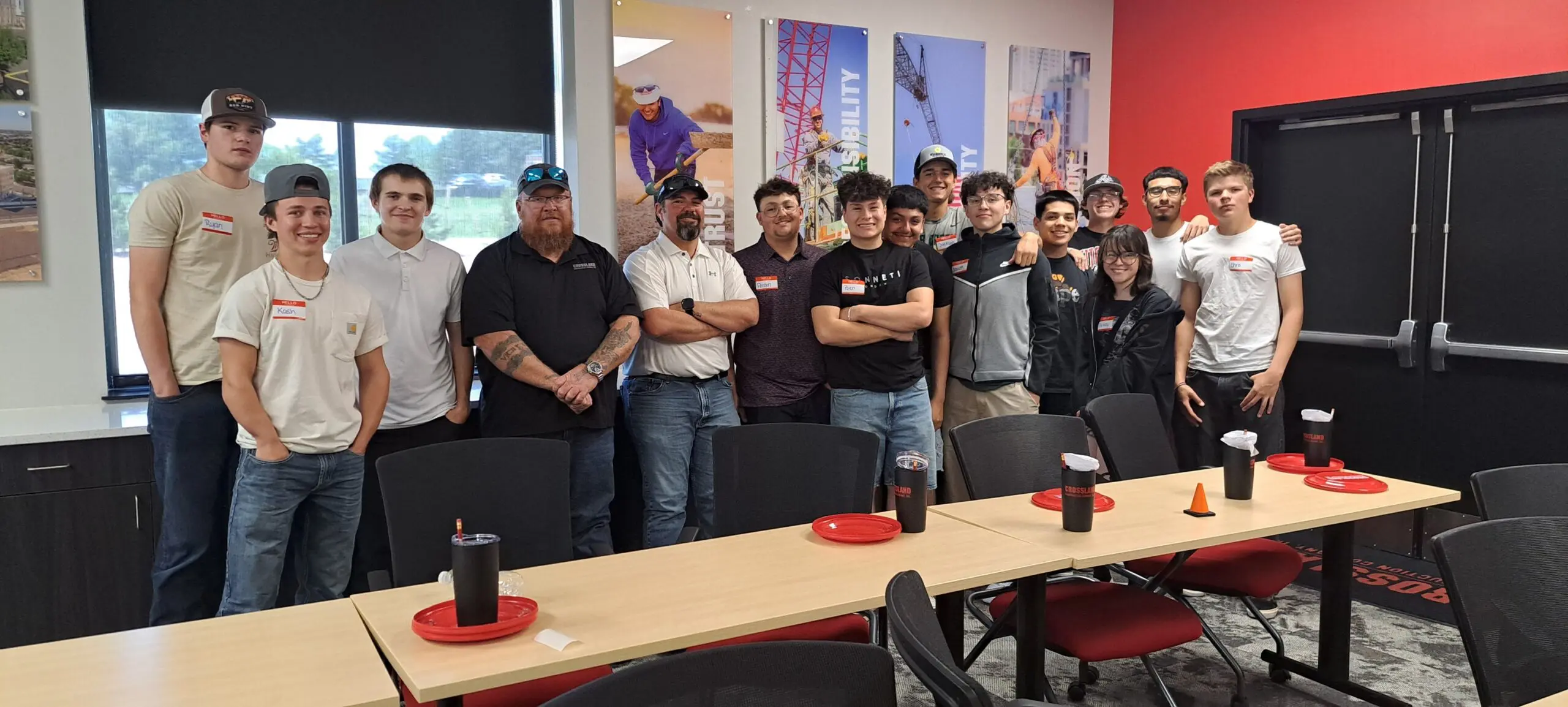 A group of people, mostly young adults, stand together and smile in a classroom or meeting room with tables, chairs, and construction-themed wall art. Some wear name tags and casual clothing; plates and napkins are on the tables.