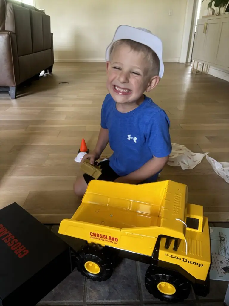 A smiling young boy in a blue shirt and white hat sits on a wooden floor, holding a toy traffic cone beside a large yellow toy dump truck labeled "CROSSLAND." Unwrapped packaging is scattered nearby.