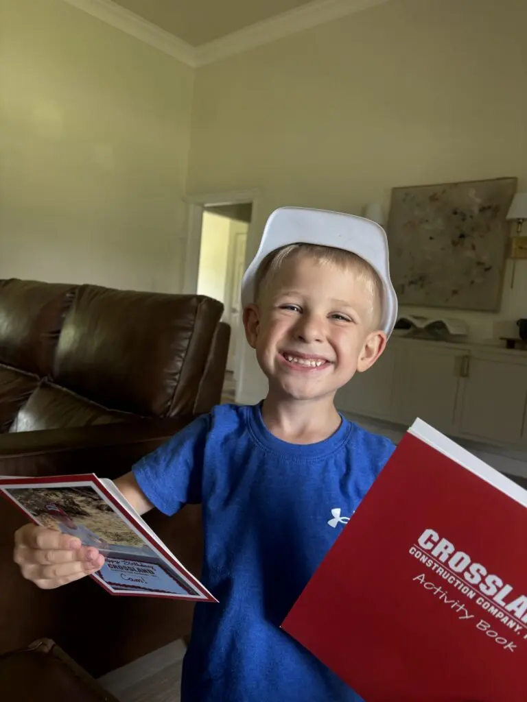 A smiling young boy in a white toy hard hat holds a red activity book and a booklet while standing in a living room with a brown leather couch and wall art in the background.