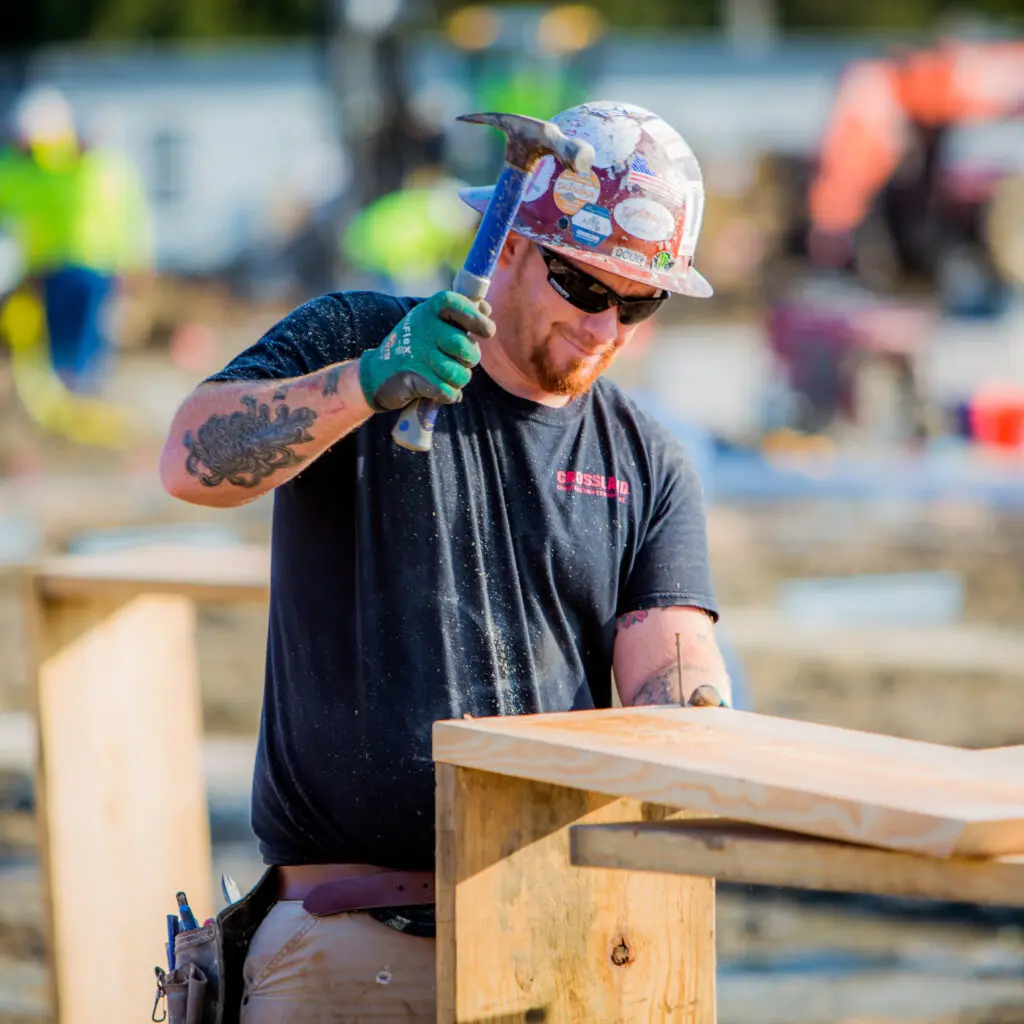 A construction worker wearing a hard hat, sunglasses, and gloves hammers a nail into a wooden board at an outdoor construction site. Other workers and equipment are blurred in the background.