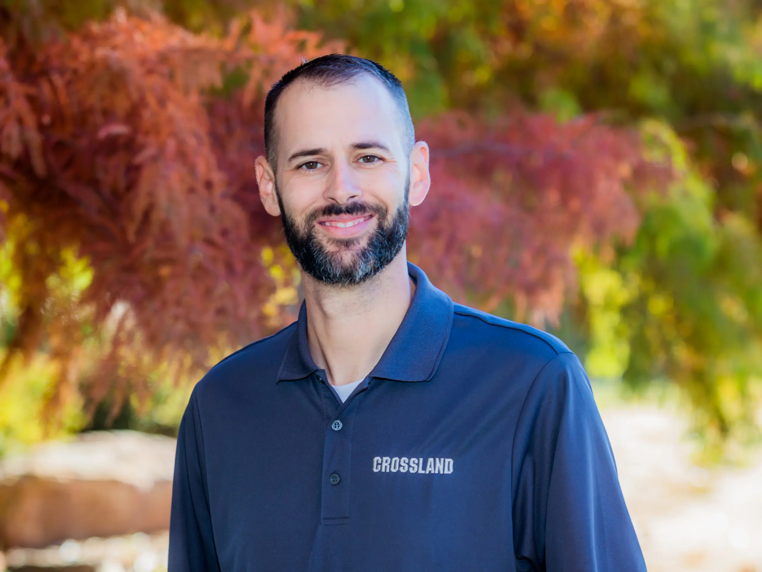 A man with short dark hair and a beard, wearing a navy blue polo shirt with the word "CROSSLAND" on it, stands outdoors in front of leafy trees with red and green foliage, smiling at the camera.