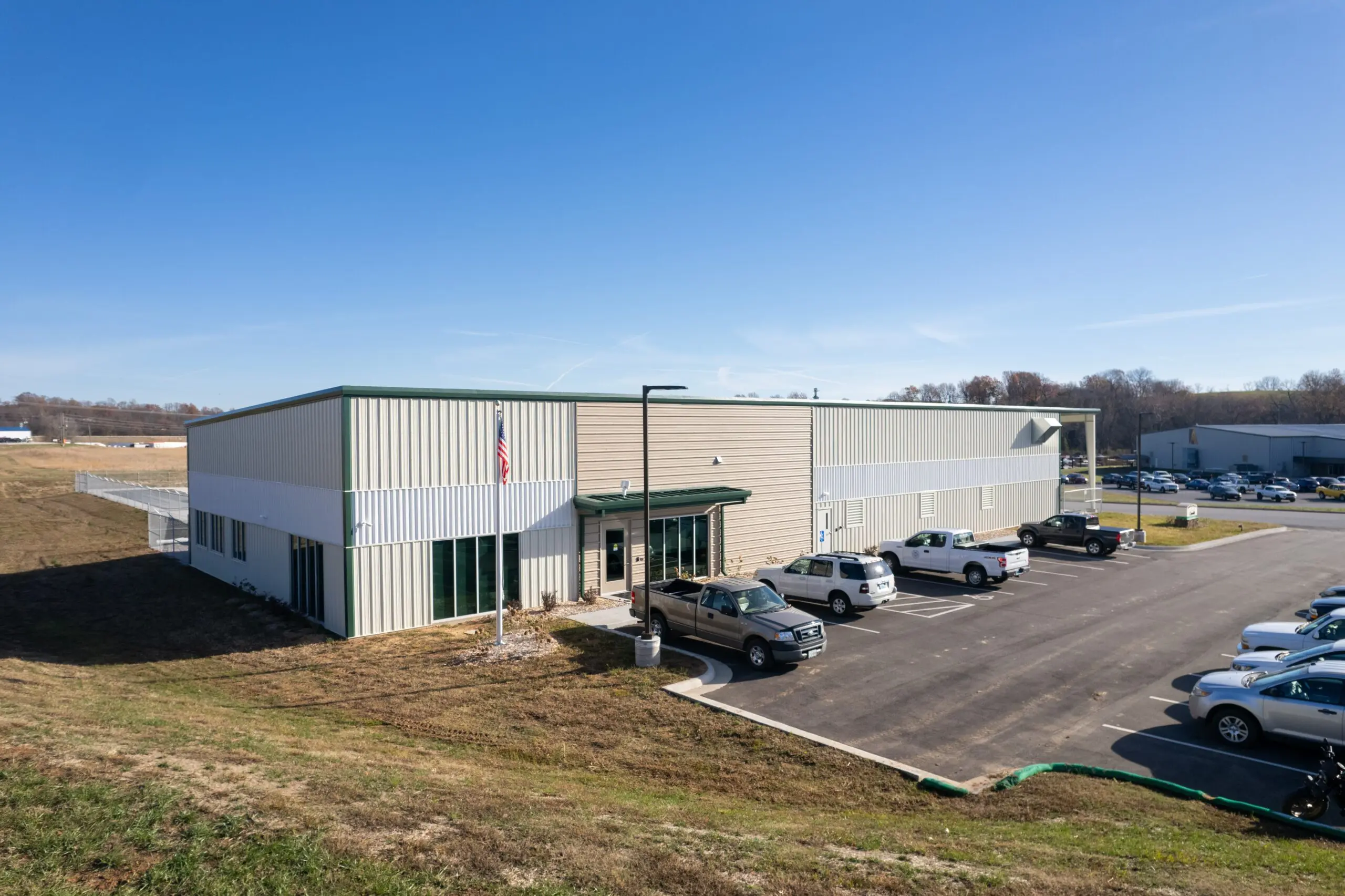 A metal industrial building with a parking lot and several vehicles parked in front, set on a grassy slope under a clear blue sky.