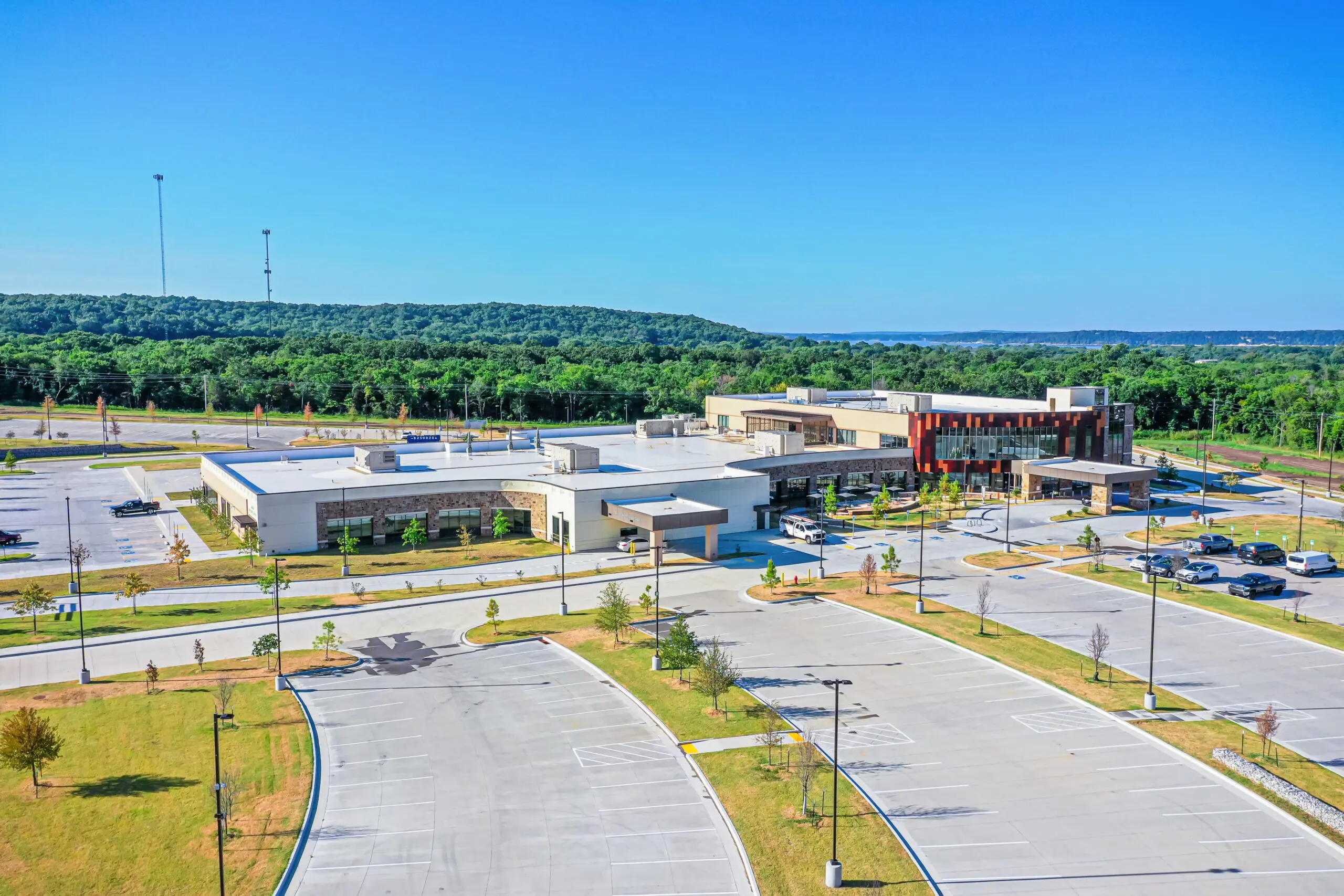 A large, modern building with surrounding parking lots and landscaped areas sits near a forested area under a clear blue sky. Few cars are parked, and the facility appears to be a hospital or medical center.