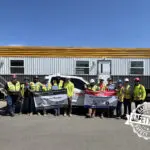 A group of construction workers in safety gear stand in front of a trailer and truck, holding banners. A large "SAFETY FIRST" graphic is stamped in the bottom right corner of the image.