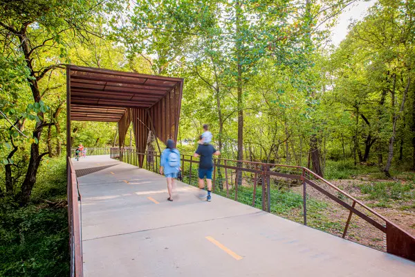 Two people walk on a paved trail through a lush, green forest. The path leads under a modern, rust-colored metal canopy structure, with more people visible in the distance enjoying the scenic, tree-filled surroundings.
