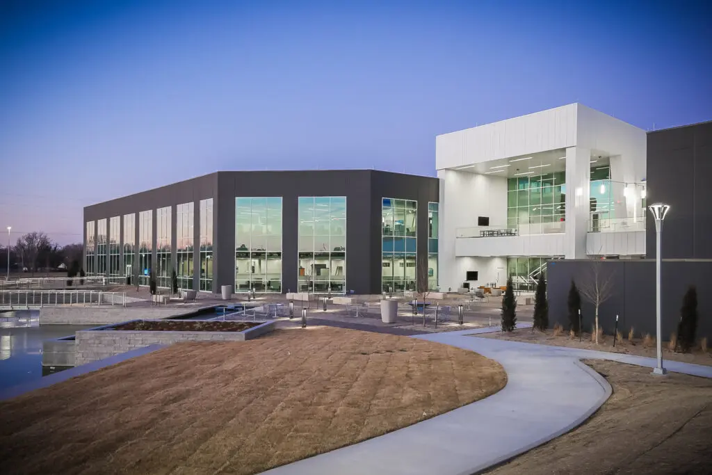 Modern building with large glass windows, surrounded by landscaped paths and grass, photographed at dusk with a clear sky in the background. Outdoor lighting is visible along the walkway.