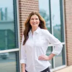 A woman with long brown hair, wearing a white button-up shirt, stands smiling with one hand on her hip in front of a building with large windows and brick walls.