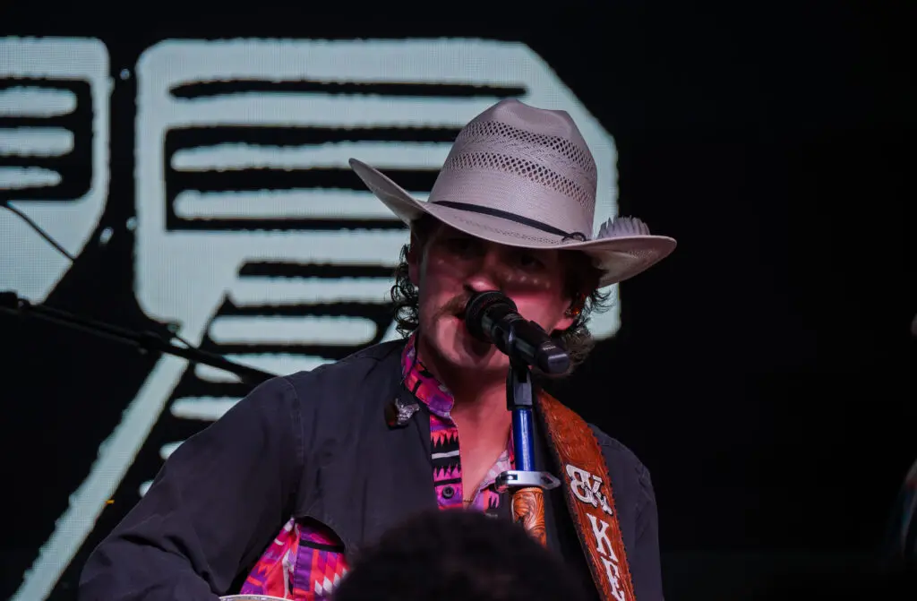 A musician wearing a cowboy hat and patterned shirt sings into a microphone on stage, holding a guitar. The background features a large, illuminated design.