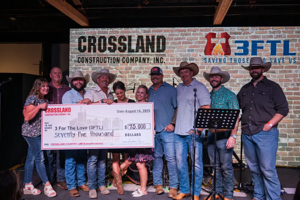 A group of people smiling on stage, holding a large $75,000 check from Crossland Construction Company Inc. to 3 For The Love (3FTL), with a brick wall and company logos in the background.