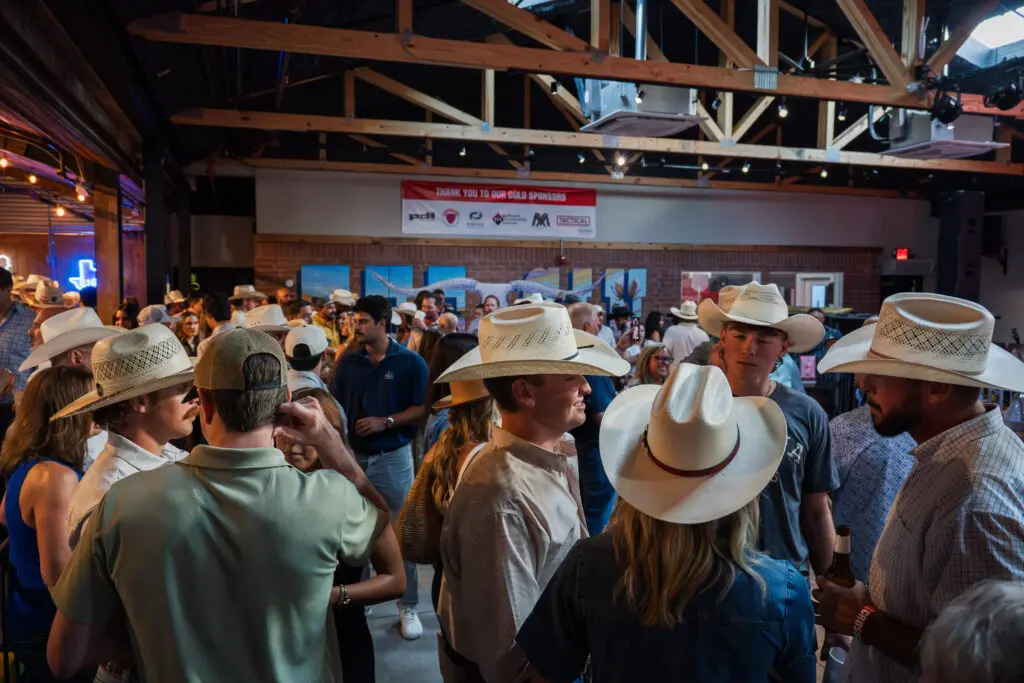 A large group of people wearing cowboy hats gathers indoors at a busy event, mingling and talking under exposed wooden beams. A banner with sponsor logos hangs on the back wall.