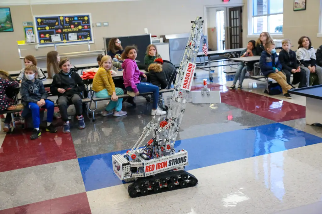 A group of children sit and watch a large remote-controlled model crane labeled "RED IRON STRONG" and "CROSSLAND" as it moves across the floor in a school cafeteria or classroom setting.