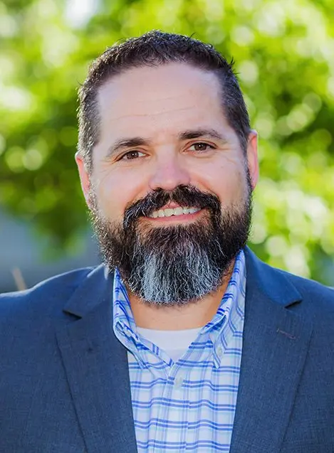 A man with a dark beard and mustache, wearing a blue suit jacket and a blue-and-white checked shirt, smiles at the camera. The background is outdoors with green foliage and sunlight.