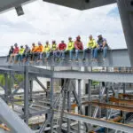 A group of construction workers wearing hard hats and safety gear sit in a row on a steel beam high above the ground at a building site, with blue sky and scaffolding visible around them.