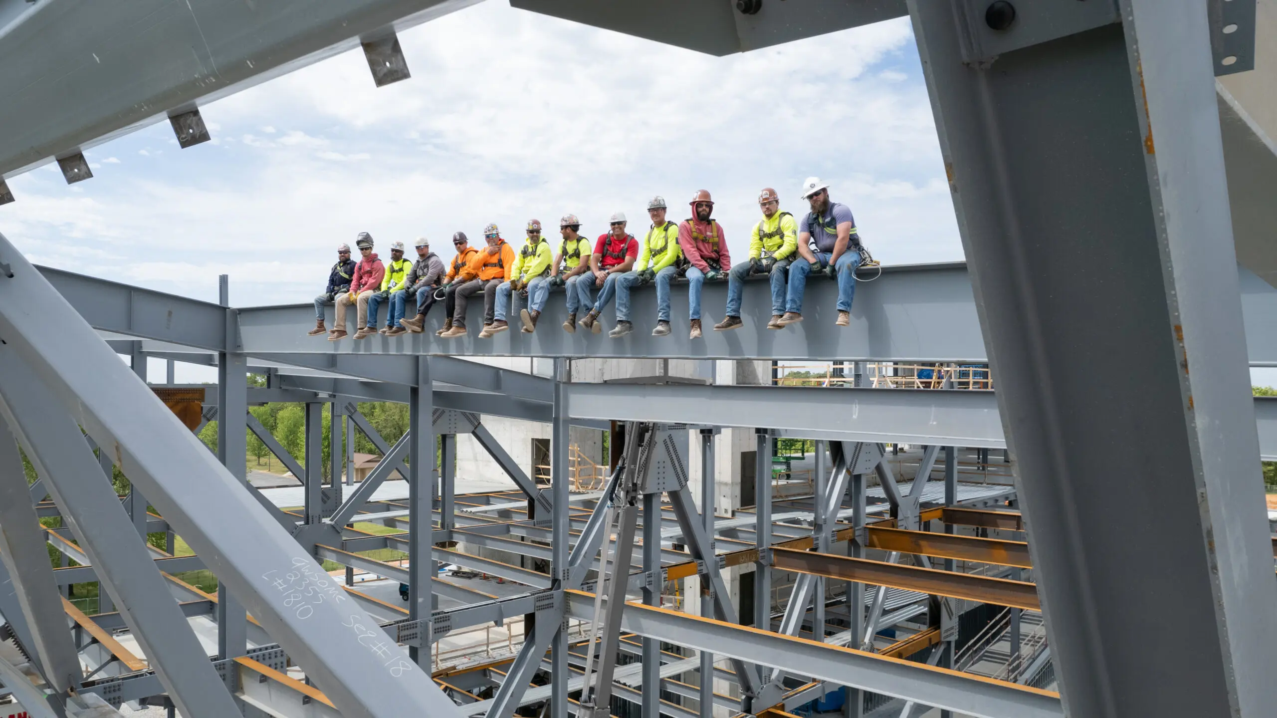 A group of construction workers wearing hard hats and safety gear sit in a row on a steel beam high above the ground at a building site, with blue sky and scaffolding visible around them.