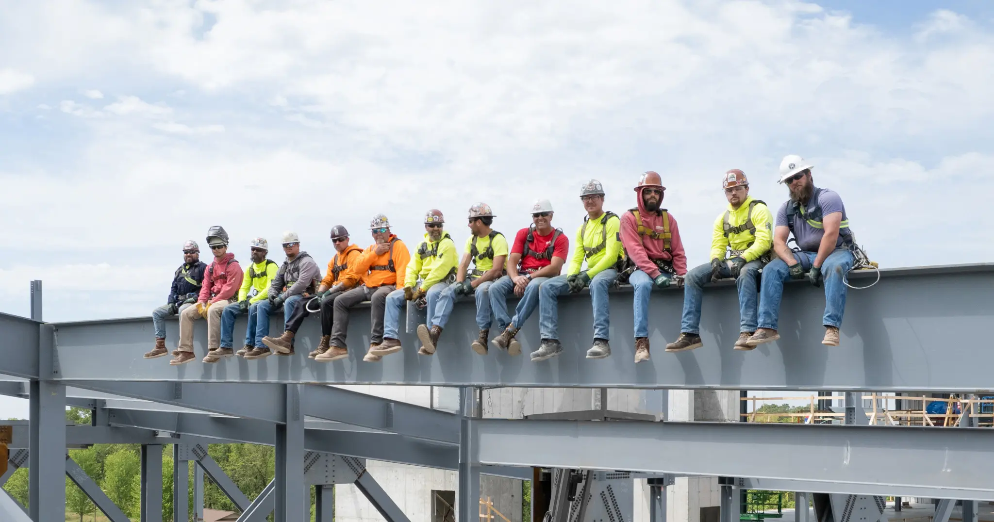 Fourteen construction workers wearing safety gear and colorful shirts sit side by side on a steel beam high above the ground at a construction site, with blue sky and trees in the background.