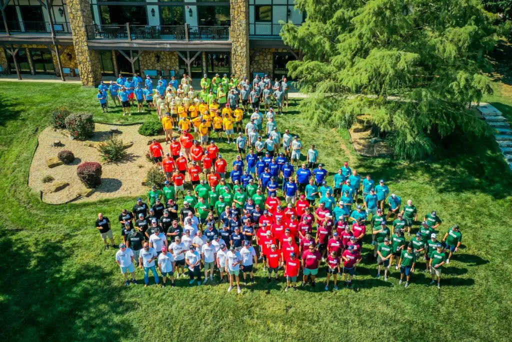 A large group of people wearing brightly colored shirts stands outside on green grass, arranged in sections by color, in front of a building with stone walls and a wooden balcony. Trees and landscaped bushes are nearby.