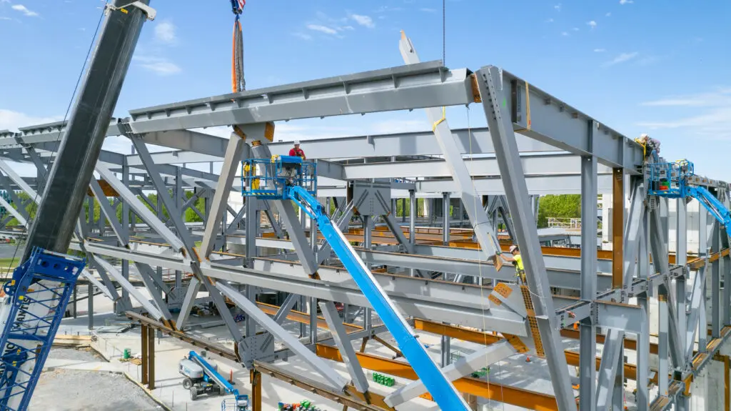 Construction workers on lifts are assembling a large steel framework for a building under a clear blue sky, with cranes and beams visible around the structure.