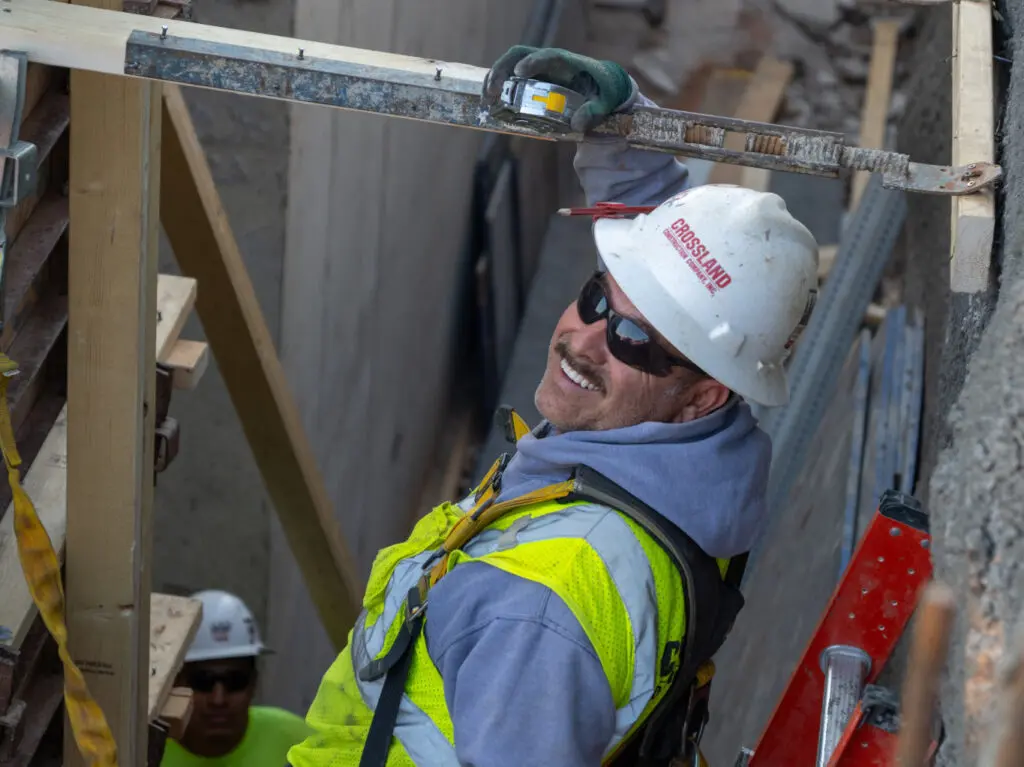 A construction worker in a white hard hat, sunglasses, and a high-visibility vest smiles while using a measuring tape at a building site. Another worker is partially visible in the background.
