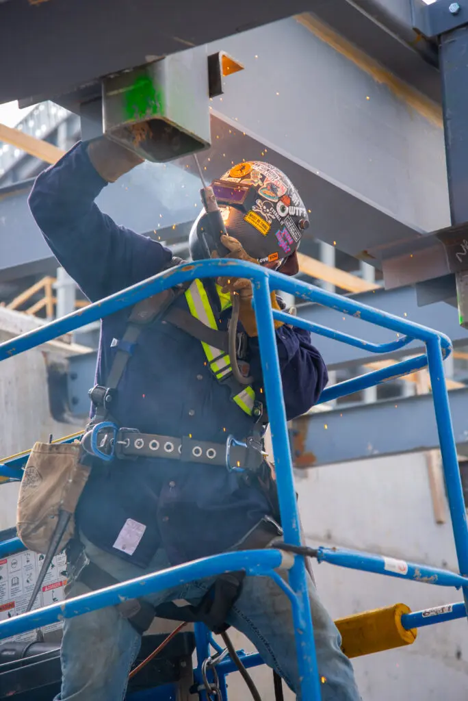 A construction worker wearing safety gear, including a helmet with stickers, harness, and gloves, uses a power tool to weld or cut metal beams on a jobsite, with sparks flying in the background.