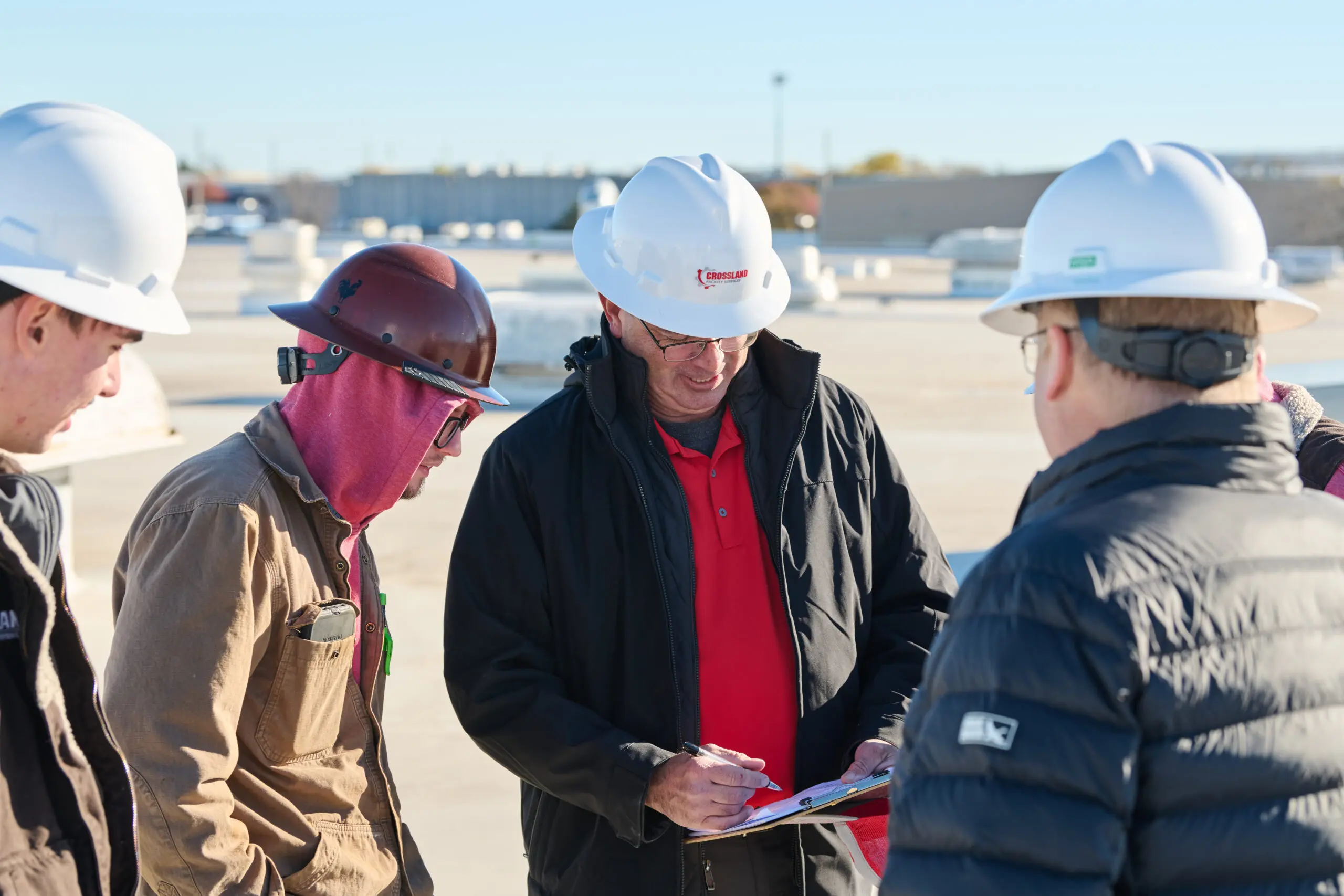 Four construction workers wearing hard hats and jackets stand together outdoors. One person in a white hard hat writes on a clipboard while others look on, appearing to discuss plans at a construction site.