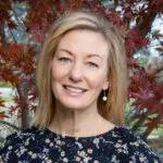 A woman with shoulder-length blonde hair and a black floral top smiles outdoors, standing in front of a tree with red leaves.
