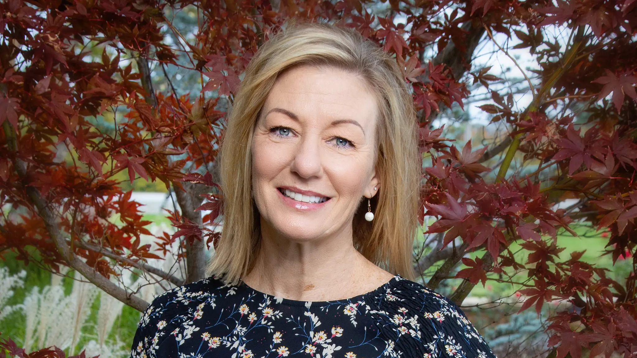A woman with shoulder-length blonde hair and a black floral top smiles outdoors, standing in front of a tree with red leaves.