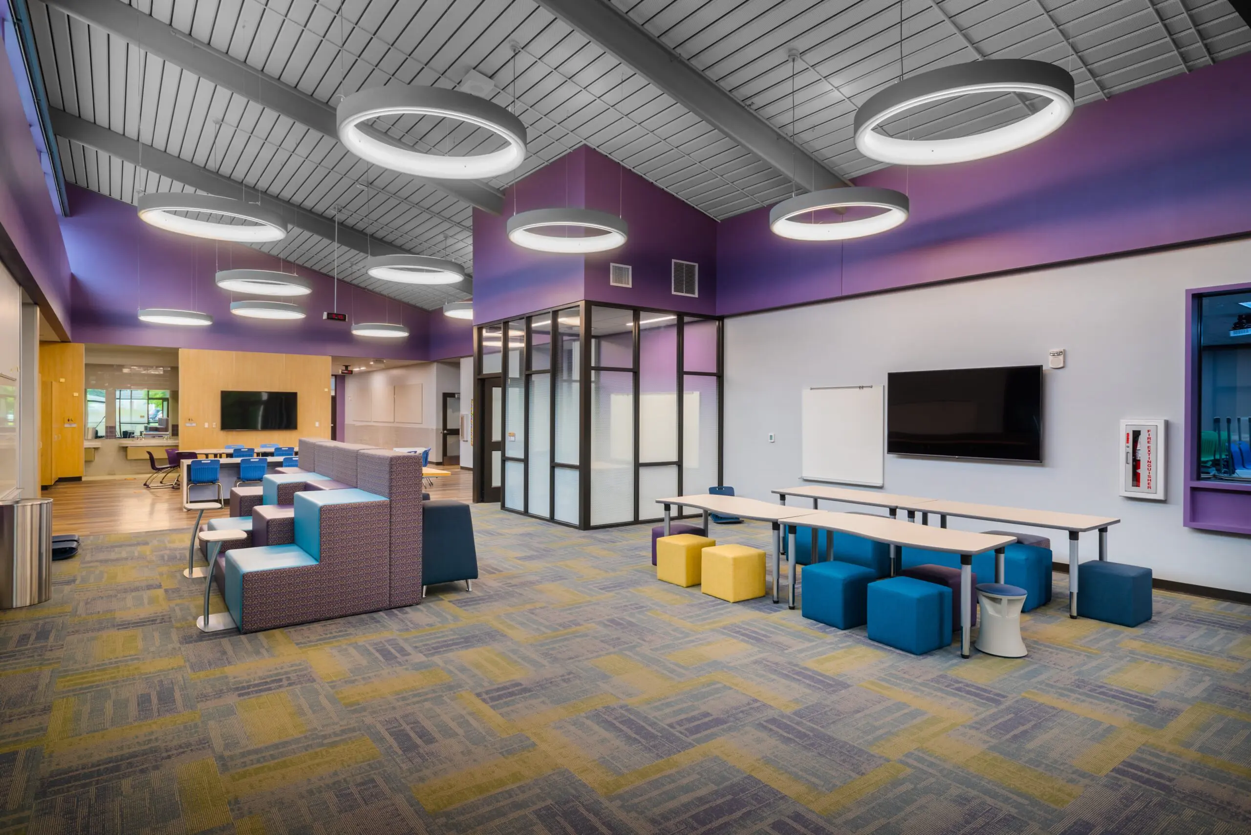 Modern, spacious lounge area with colorful seating, geometric furniture, circular ceiling lights, glass-walled rooms, and a wall-mounted monitor. The space features a mix of tables, chairs, and soft seating on patterned carpet.