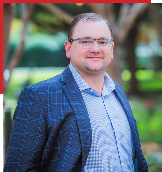 A man in glasses and a blue plaid suit jacket stands outdoors, smiling slightly. The background is blurred greenery with trees, and the image has a partial red border on the top and left sides.