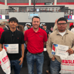 Three men stand indoors at an event. The man in the center, wearing a red polo, is flanked by two young men holding certificates and gift bags. People and tables are visible in the background.