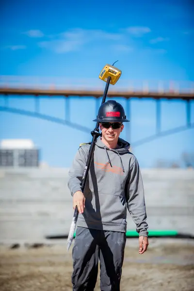 A construction worker in a gray hoodie and black hard hat holds surveying equipment over his shoulder, smiling at a construction site with blue sky and a partially built structure in the background.