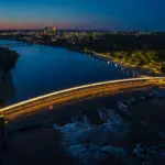Aerial view of a brightly lit pedestrian bridge curving over a river at dusk, with city lights and buildings visible in the background and gentle water currents below the bridge.