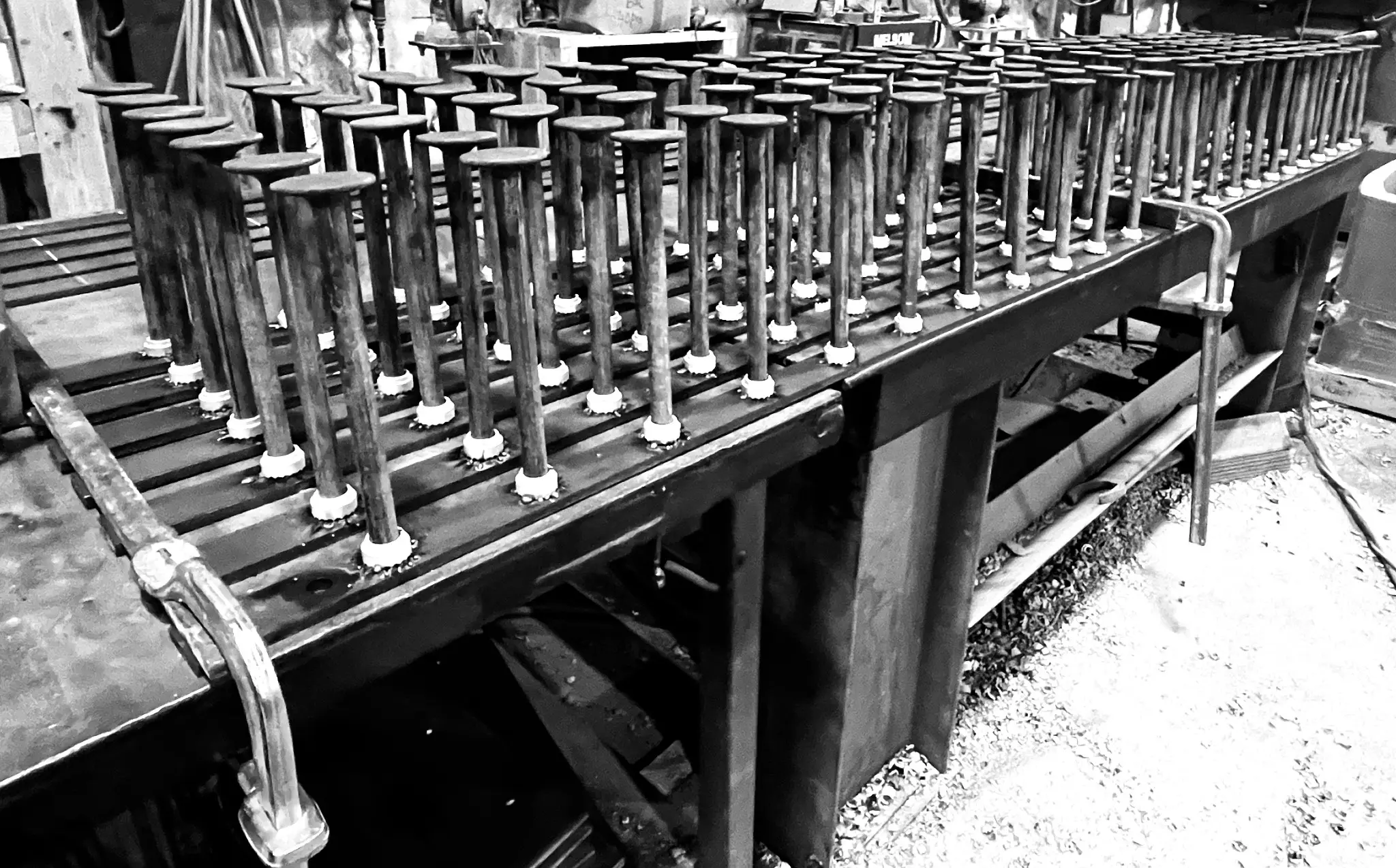 Black and white image of a metal industrial table with numerous vertical cylindrical rods evenly spaced in rows, likely part of a factory or workshop setting. The background shows various equipment and tools.