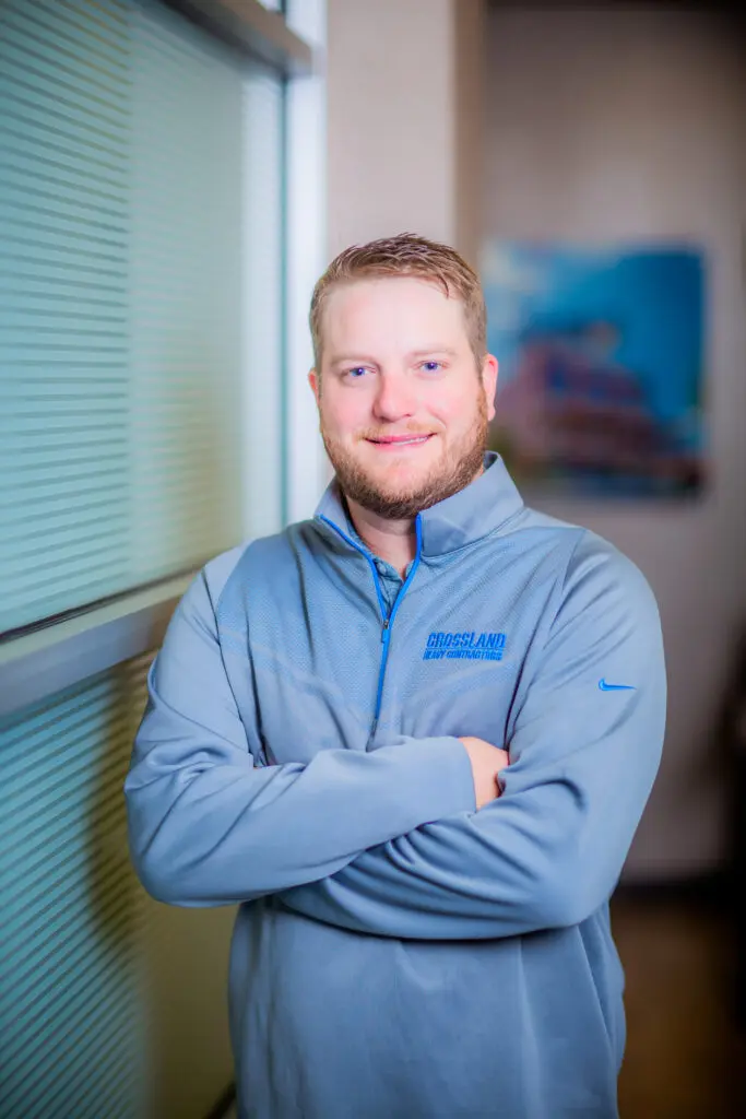 A man with short blonde hair and a beard stands indoors with arms crossed, wearing a light blue quarter-zip pullover. He is smiling and standing next to a window with blinds.