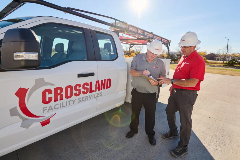 Two men in hard hats and uniforms stand next to a Crossland Facility Services truck on a sunny day, reviewing paperwork or a clipboard together on a paved surface.