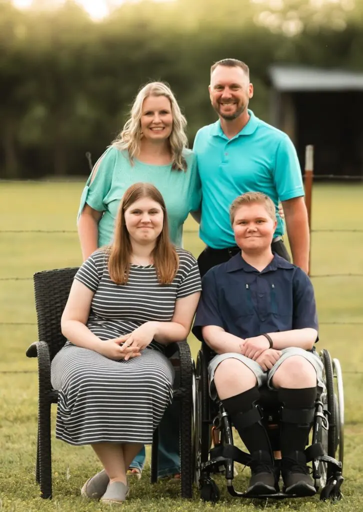 A smiling family of four poses outdoors. Two adults stand behind two teens; one teen sits on a chair, and the other uses a wheelchair. They are in front of a wire fence with green grass and trees in the background.