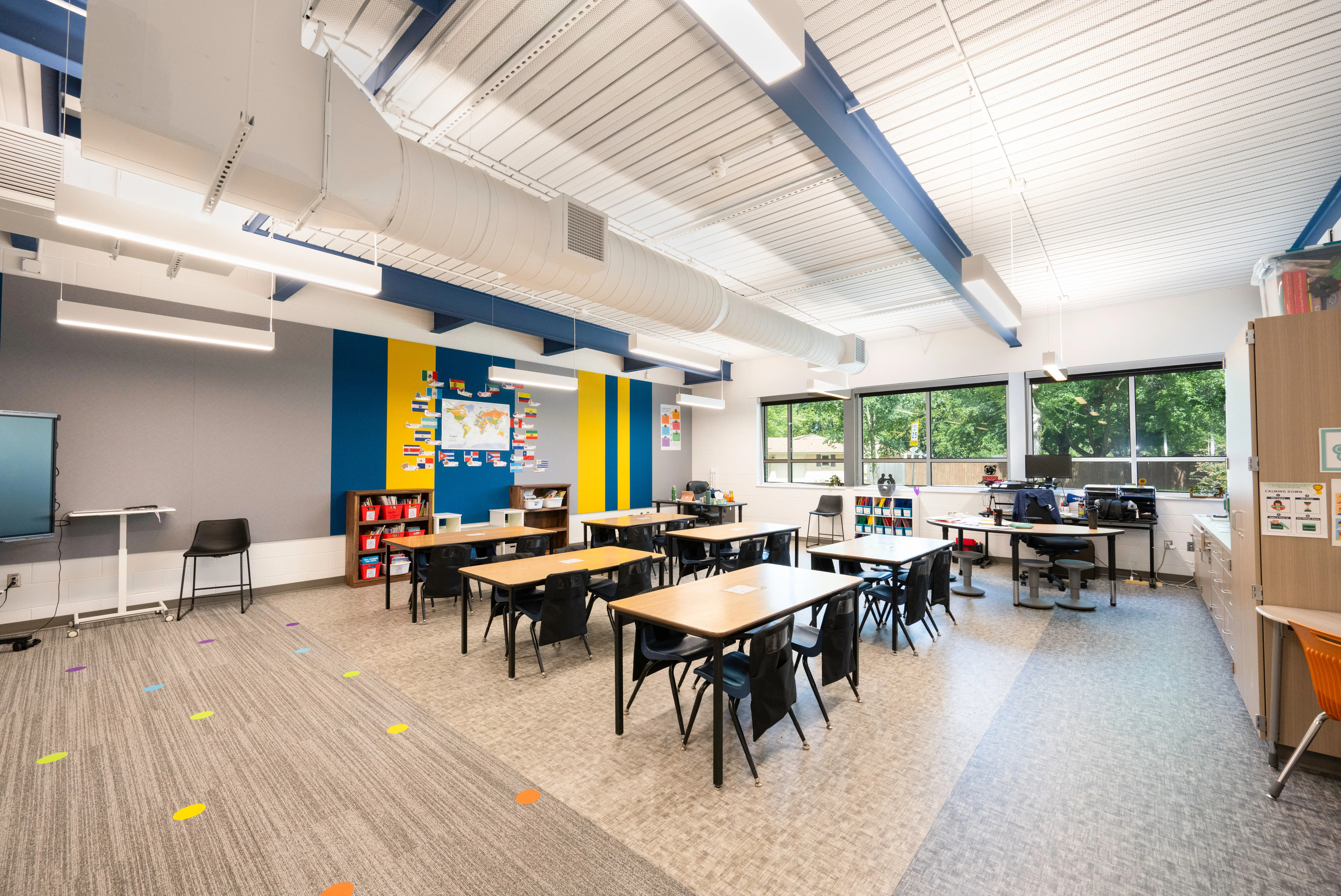 A bright, modern classroom with several tables and black chairs, colorful wall decor, a world map, large windows letting in natural light, and shelves with books and educational materials.