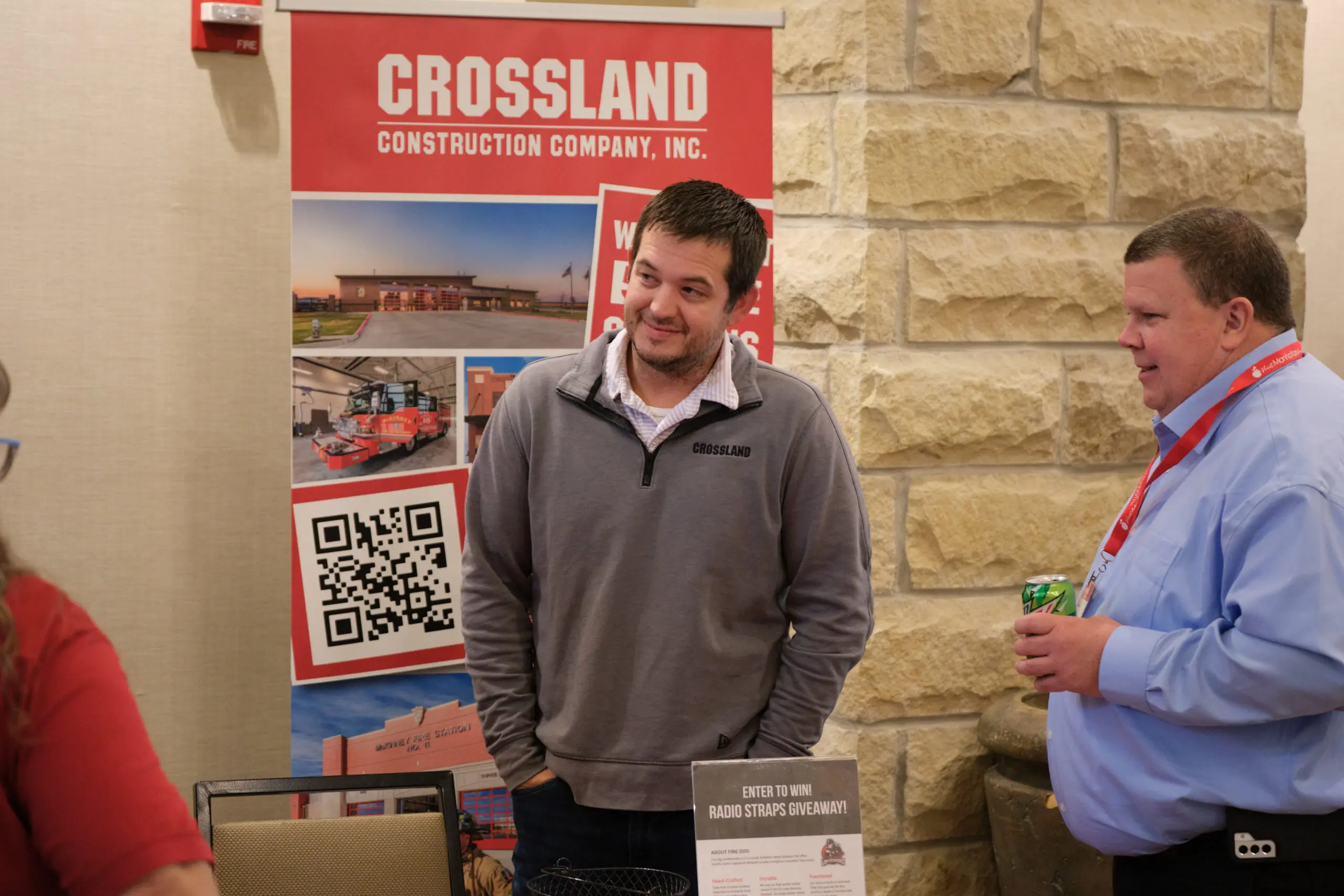Two men stand talking at a booth for Crossland Construction Company, Inc. One man wears a gray pullover with the company logo, and behind them is a promotional banner with images, text, and a QR code.
