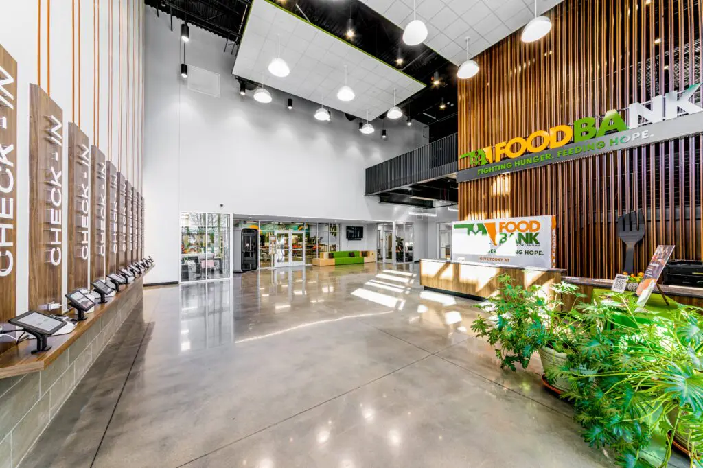 Spacious, modern food bank lobby with tall ceilings, polished floors, wooden slat walls, check-in stations on the left, and a large “Food Bank” sign above the main desk. Plants and natural light add warmth to the space.