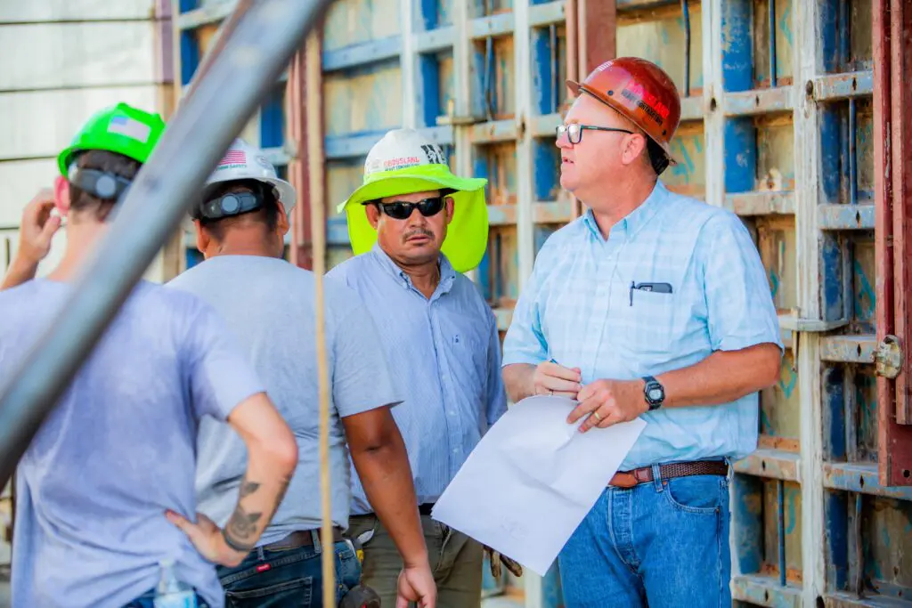 A group of construction workers wearing helmets and safety gear stand near a wall structure. One man in a red helmet and glasses holds papers and appears to be giving instructions to the others.
