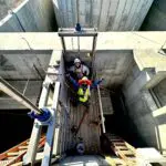 Two construction workers wearing safety gear stand in a concrete structure under bright sunlight, looking up and gesturing. Various construction tools and equipment surround them within the worksite.