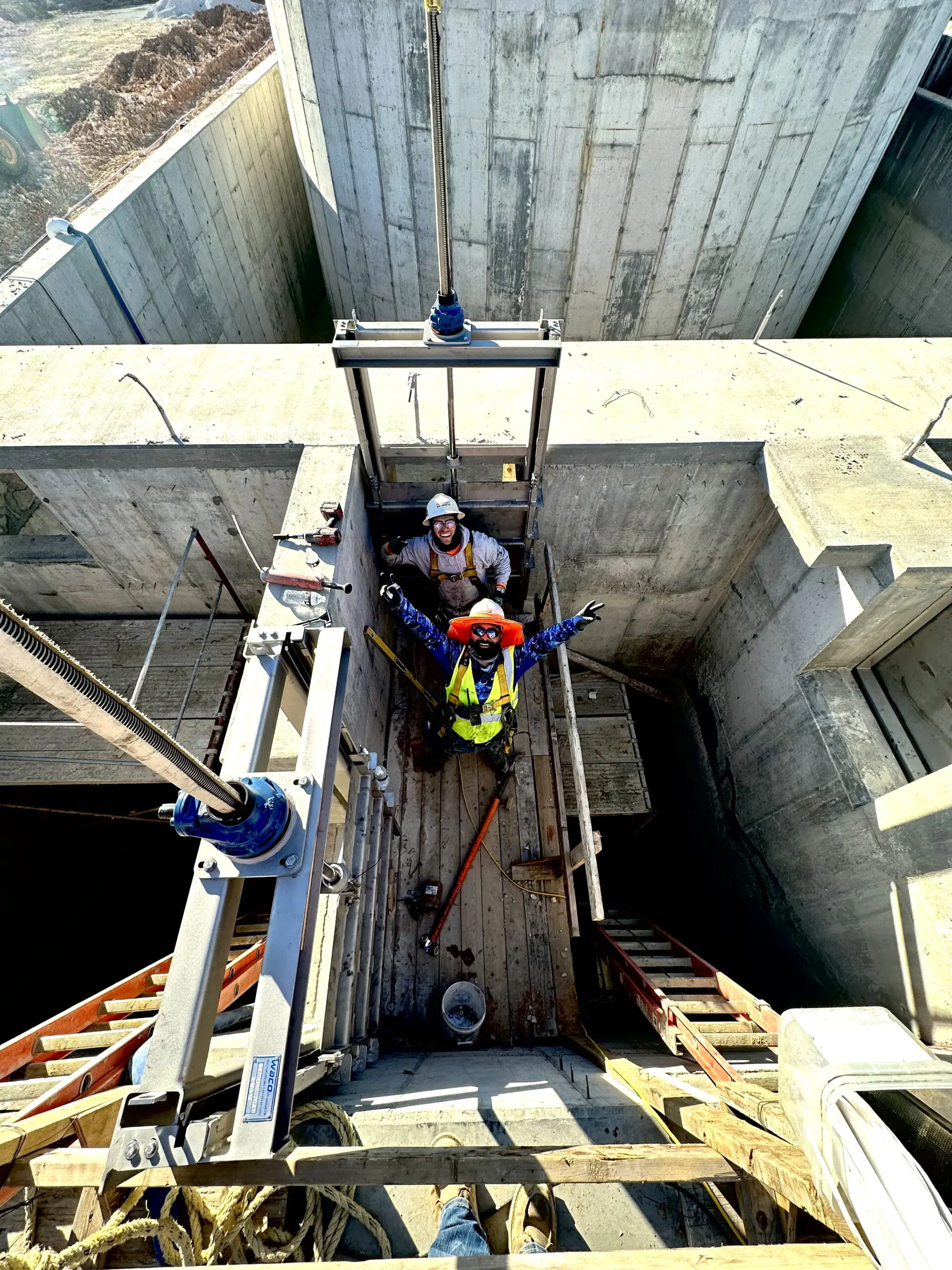Two construction workers wearing safety gear stand in a concrete structure under bright sunlight, looking up and gesturing. Various construction tools and equipment surround them within the worksite.