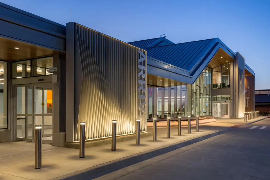 Modern airport terminal exterior at dusk with illuminated bollards, large glass windows, and a sign that reads "ARRIVE" next to the entrance. The sky is clear, and the building features contemporary design elements.