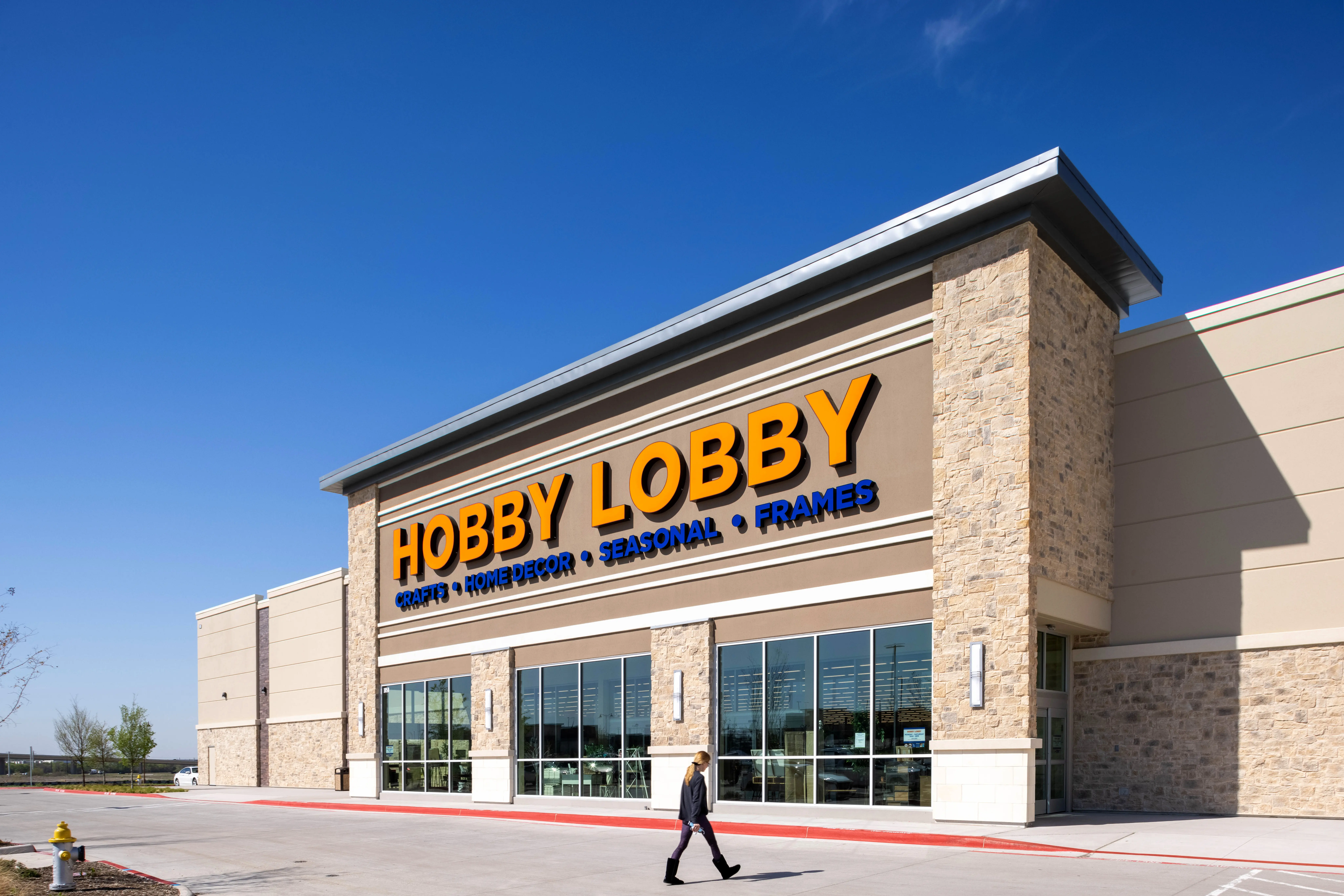 A person walks in front of a Hobby Lobby store with large orange and blue signage on a sunny day. The building has stone accents and large windows.