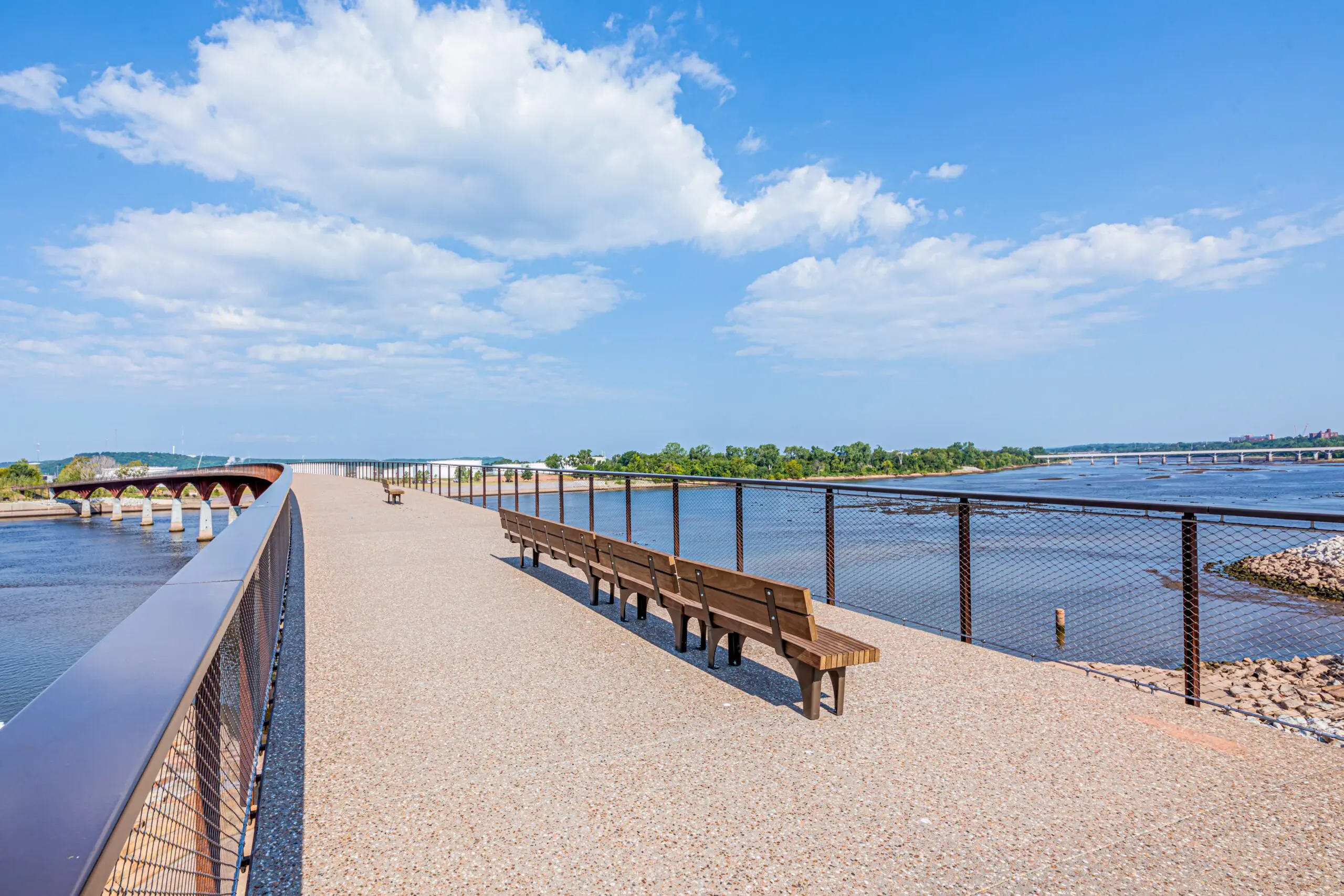 A wide pedestrian bridge with benches crosses over a river on a sunny day, with blue sky, scattered clouds, and trees in the background.