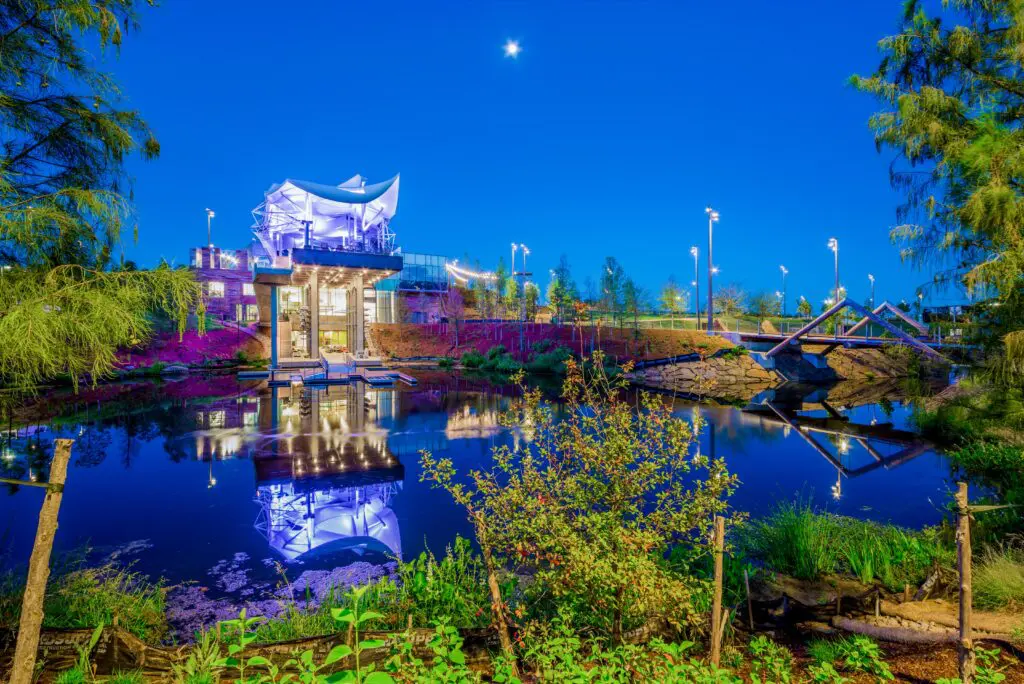 A modern, illuminated structure stands beside a reflective pond at dusk, surrounded by trees and greenery, with a clear blue sky and bright star overhead. Lights and a small bridge enhance the peaceful scene.