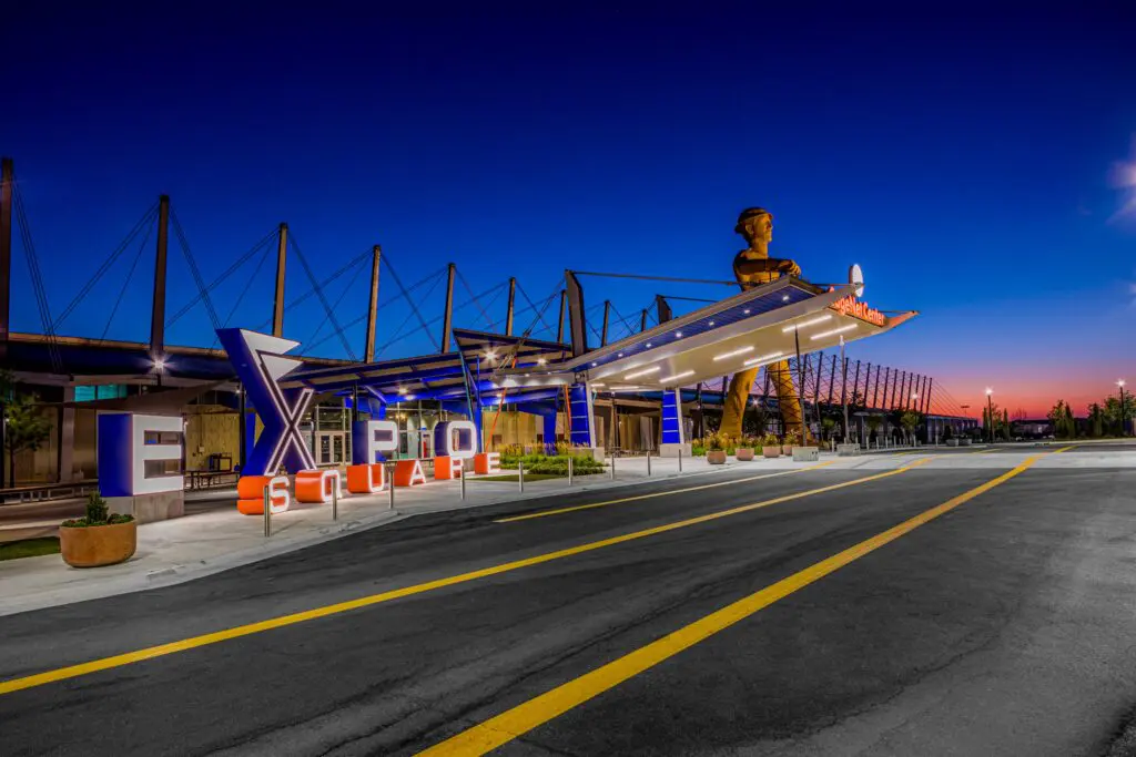 The entrance to Expo Square in Tulsa, Oklahoma is lit up at dusk, featuring a large illuminated sign and the iconic Golden Driller statue in the background against a deep blue and orange sky.