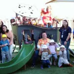 A group of adults and children pose and smile together around a playground structure with a slide on a sunny day. Some children wear hats and costumes, and everyone looks happy.